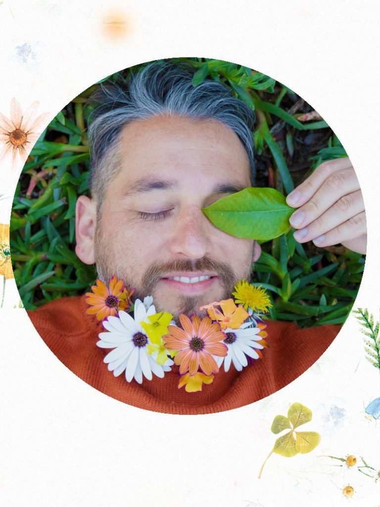 Ein Mann mit Blumen und Blättern um das Gesicht, entspannt auf grünem Gras.